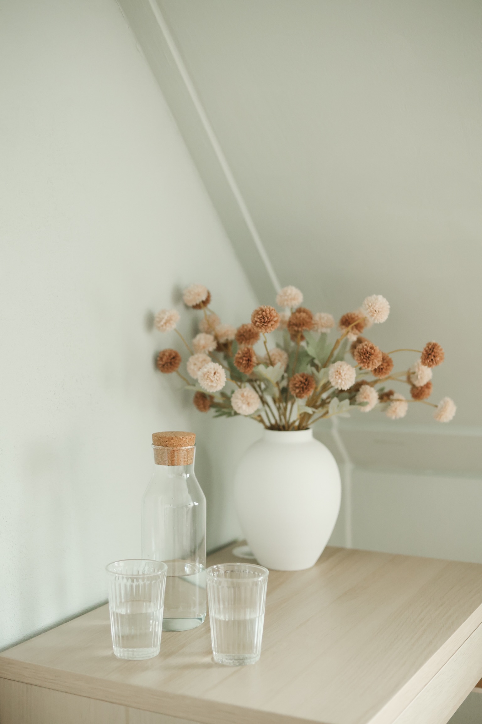 Een vaas met gedroogde bloemen, een karaf en twee waterglazen op een plank onder het schuine dak