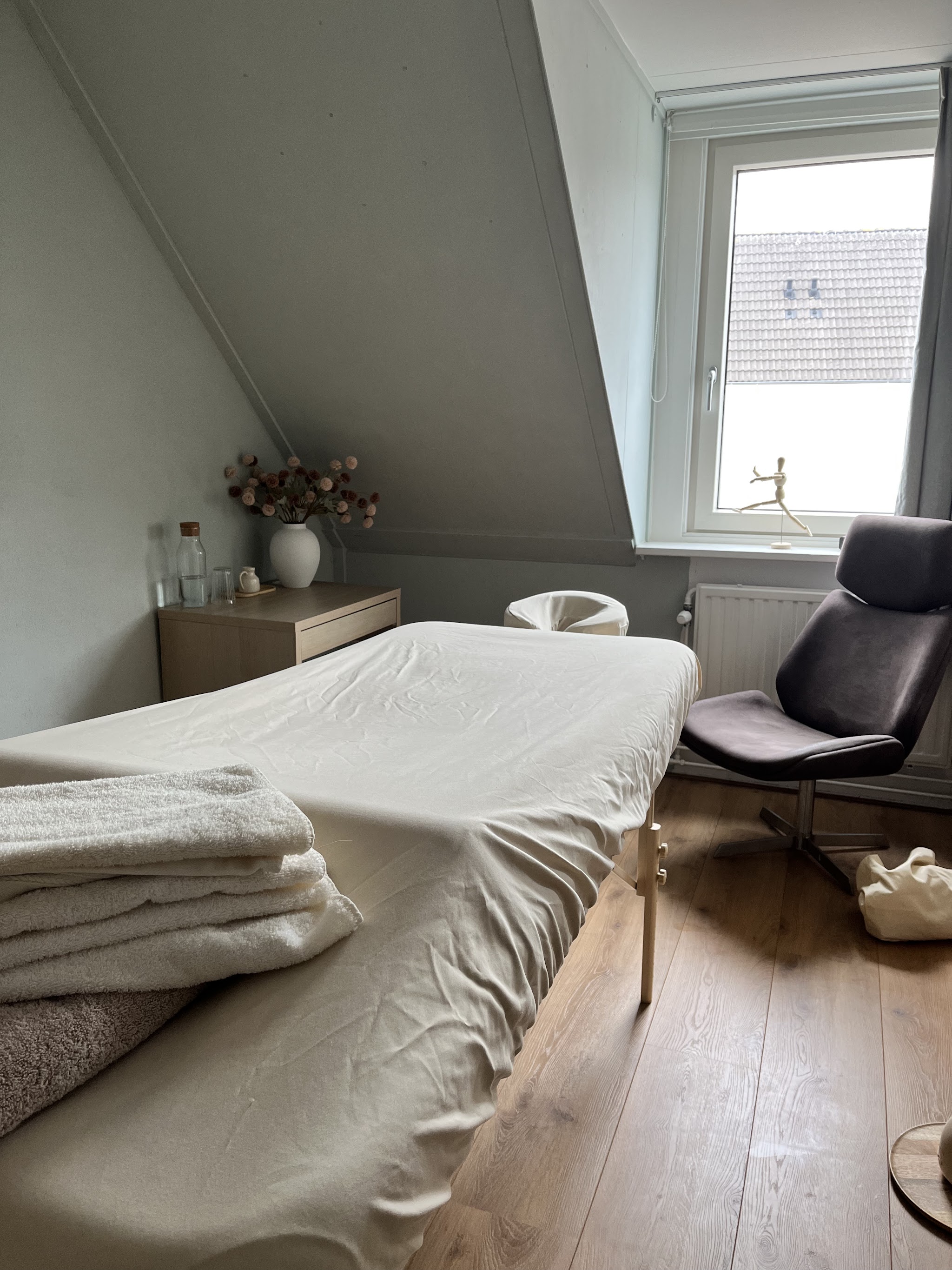De behandelkamer onder het schuine dak: de massagetafel met linnen en handdoeken, een fauteuil bij het raam
